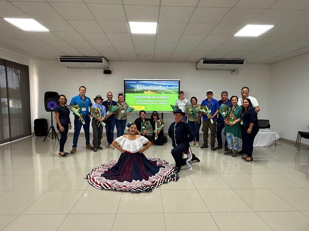 Un grupo de personas de pie sosteniendo ramos de flores, al frente una mujer y un hombre con vestido y traje típico de folclor costarricense